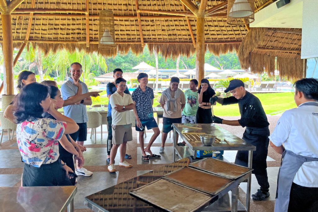 Guests at Atmosphere Resorts & Spa enjoy an interactive cooking demonstration with Chef Jun Tubog under a thatched pavilion, sharing laughter and learning about Filipino cuisine.