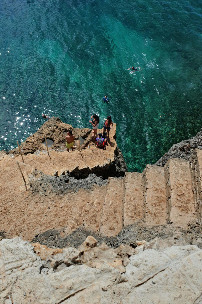People standing on rocky steps leading down to clear turquoise water at a popular swimming spot in Siquijor.