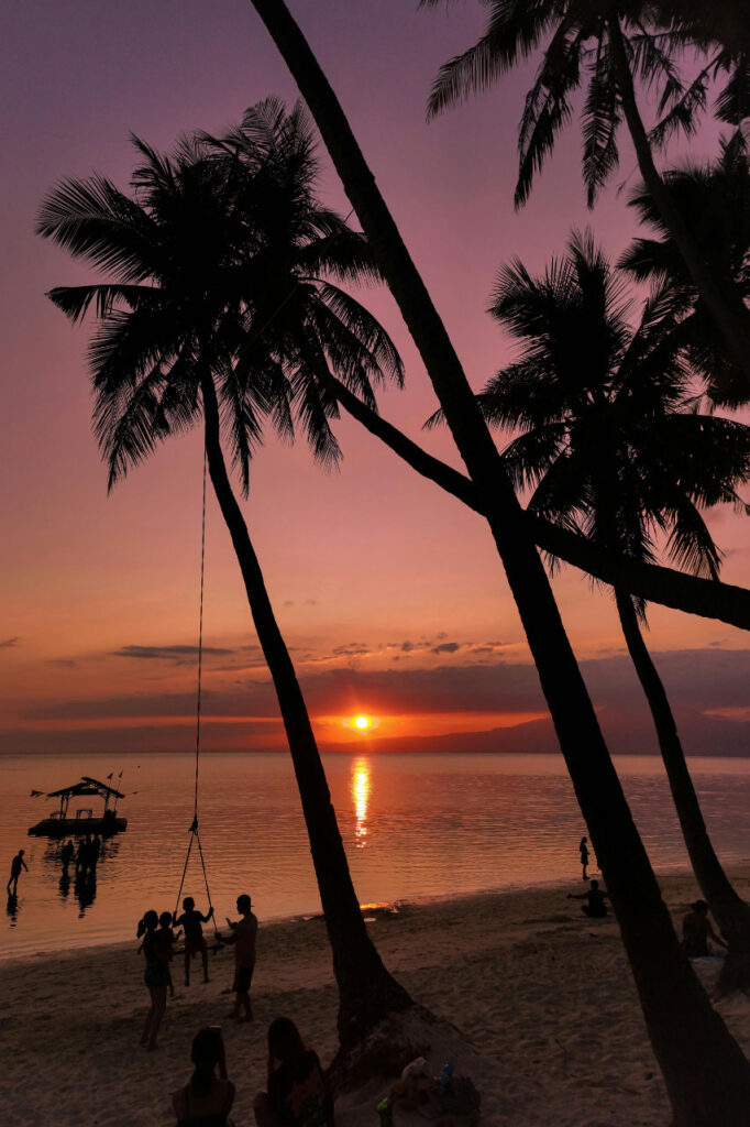 Sunset at Paliton Beach in Siquijor with silhouetted palm trees, people on the shore, and a boat on the calm sea.