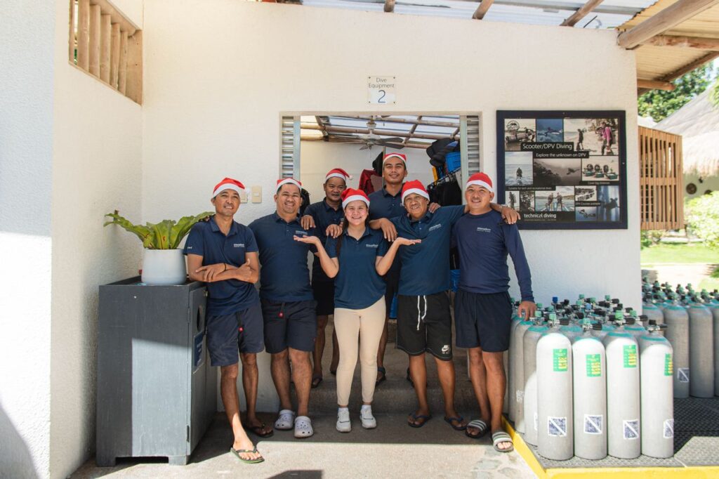 Atmosphere Resorts & Spa dive team wearing Santa hats, standing together at the dive center beside scuba tanks.