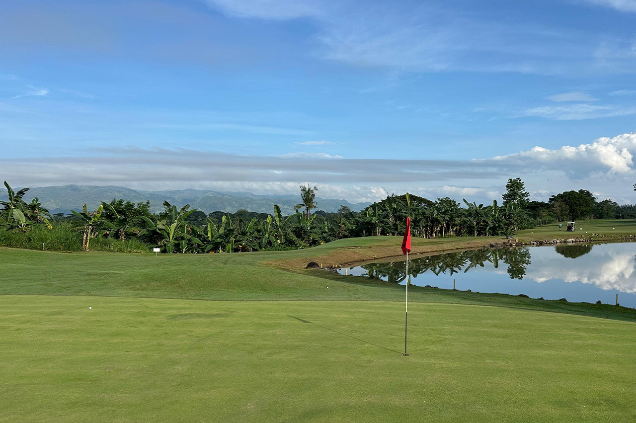 Putting green with flag beside a water hazard at Negros International Golf & Country Club, surrounded by tropical landscape.