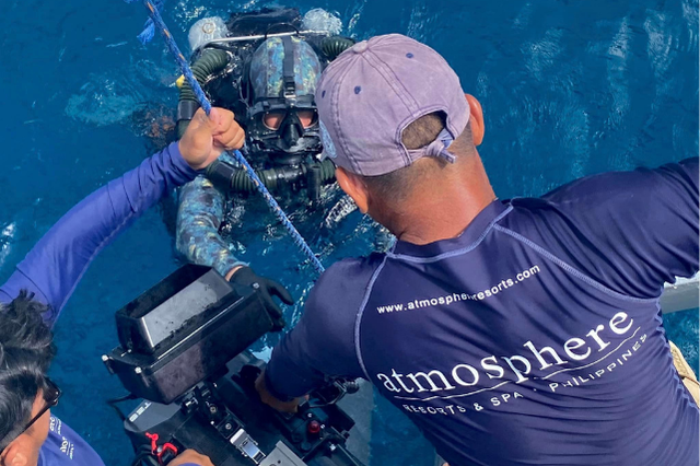 An Atmosphere Resorts & Spa crew member assists a diver in technical gear while underwater filming equipment is prepared beside a dive boat.