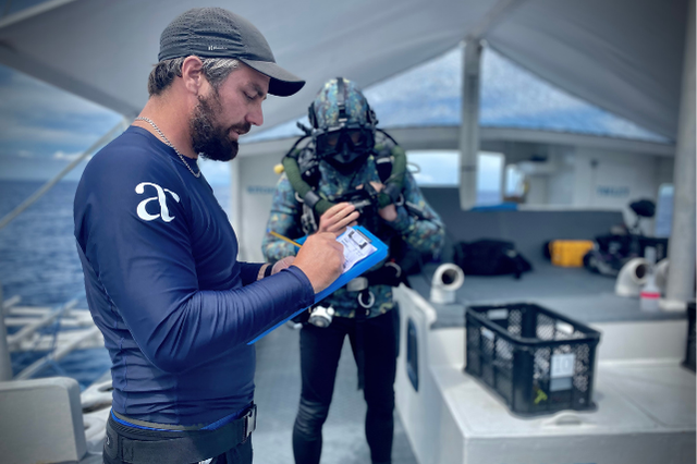 A technical diving instructor at Atmosphere Resorts & Spa reviews dive information on a tablet while a diver in full gear prepares in the background on a dive boat.