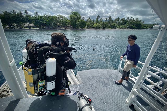 A diver wearing advanced rebreather equipment prepares to enter the water from a dive boat while a crew member stands nearby in Dauin, Negros Oriental.