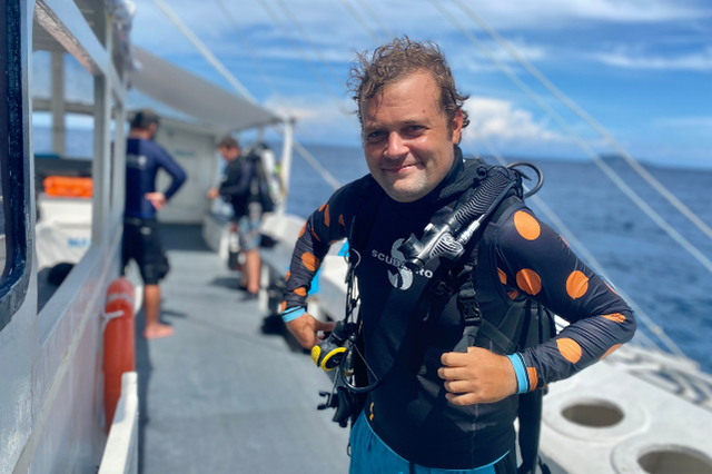 Marine biologist Daniel Geary stands aboard a dive boat in scuba gear, wearing a black rash guard with orange polka dots during a dive expedition in Dauin.