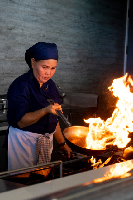 A chef at Atmosphere Resorts & Spa prepares a dish over an open flame in the resort’s kitchen, showcasing professional culinary techniques.