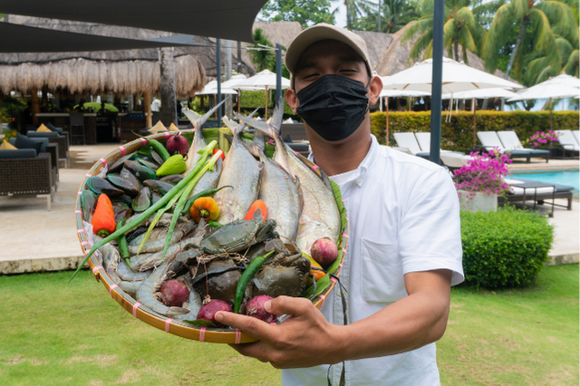 A resort team member presents a basket of freshly caught seafood and locally sourced vegetables, including fish, crab, onions, chilies, and herbs, at Atmosphere Resorts & Spa.
